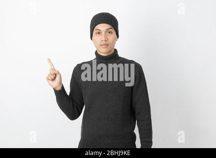 Giovane uomo asiatico bello che indossa maglione grigio e beanie che punta al lato con le dita per presentare un prodotto o un'idea su sfondo bianco. Foto Stock