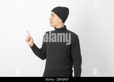 Giovane uomo asiatico bello che indossa maglione grigio e beanie che punta al lato con le dita per presentare un prodotto o un'idea su sfondo bianco. Foto Stock