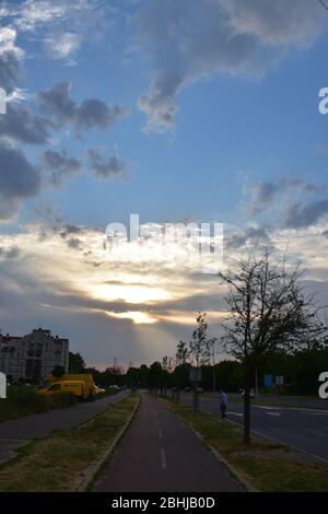 Paesaggio urbano con edifici in lontananza e infrastrutture di trasporto. Nel cielo, il sole splende dietro le nuvole Foto Stock