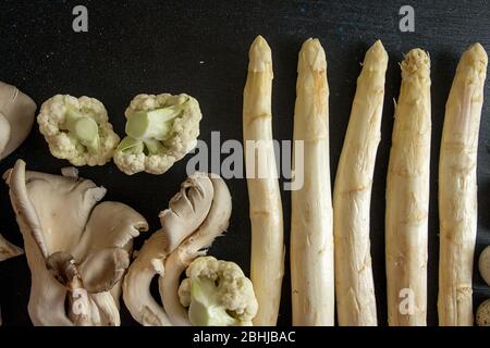 Ingredienti crudi preparati per la cottura su sfondo nero: Asparagi bianchi, funghi, cavolfiore. Overhead. Vista dall'alto. Foto Stock