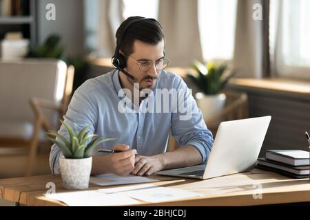 Uomo concentrato che indossa le cuffie e scrive appunti, studiando online Foto Stock