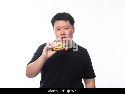 Giovane uomo grasso asiatico in polo nero mangiare hamburger con faccia felice isolato su sfondo bianco. Foto Stock