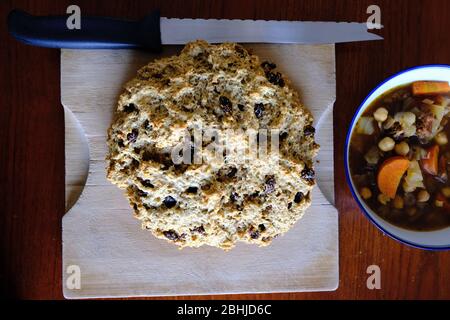 Pane fresco di soda irlandese (con uvetta) su un tagliere di legno, accanto a una ciotola blu di fagioli e stufato di verdure. Era una cena buona! Foto Stock