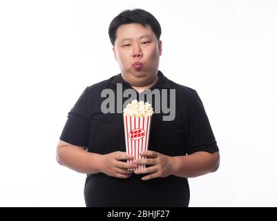 Divertente giovane asiatico grasso uomo in polo nero camicia tenendo popcorn isolato su sfondo bianco. Foto Stock