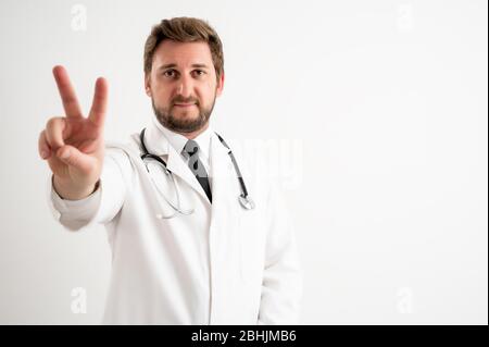 Ritratto di medico maschile con stetoscopio in uniforme medica mostrando segno di vittoria posando su un fondo bianco isolato Foto Stock