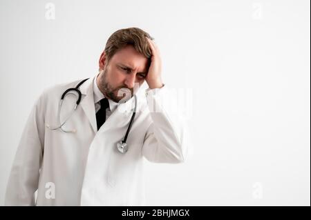 Ritratto di medico maschile con stetoscopio in uniforme medica con braccia aperte guardando in su, ha mal di testa in posa su un bianco isolato sfondo Foto Stock