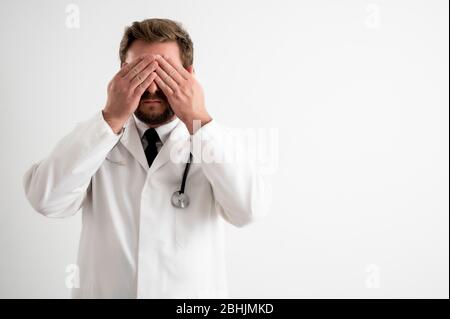 Ritratto di medico maschile con stetoscopio in uniforme medica coprente occhio come cieco concetto posando su un bianco isolato sfondo. Foto Stock