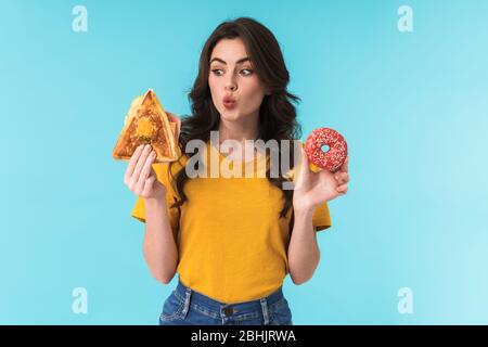 Immagine di sorpresa giovane bella donna posa isolata su sfondo blu parete tenendo sandwich e ciambella. Foto Stock