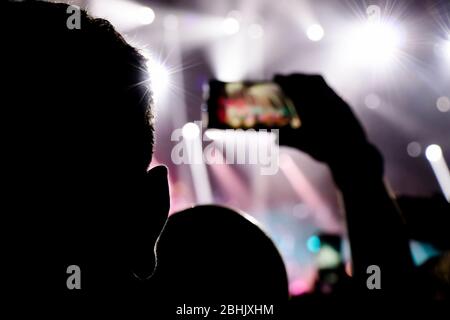 Silhouette di un orecchio mans e di un altro mans mano che spara un concerto con cellulare con effetti luce sullo sfondo, da dietro Foto Stock