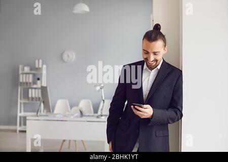 Un uomo d'affari in una giacca nera in piedi legge un messaggio in un telefono cellulare in un ufficio bianco. Foto Stock