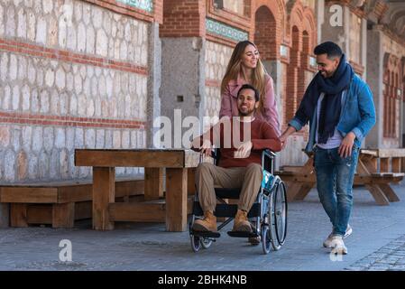 Un giovane latino-americano paralizzato in carrozzina accompagnato da una giovane ragazza caucasica e latino-americana che passeggiando felicemente per la strada Foto Stock