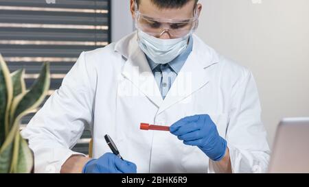Mano di un medico che tiene un flaconcino di campione di sangue e scrive ai file. Foto Stock