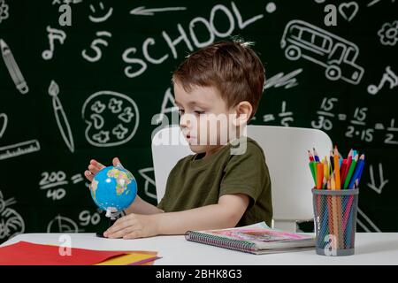 Carino ragazzo intelligente è seduto a una scrivania con globo in mano sullo sfondo con lavagna . Pronto per la scuola. Ritorno a scuola. Foto Stock