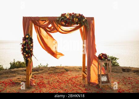 Arco in legno quadrato per matrimoni all'aperto al tramonto. Fiori rossi di rosa e stoffa appesa. Incredibile luce solare calda. Foto Stock