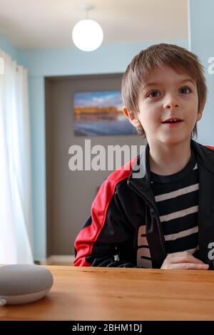 Un giovane ragazzo, di sei anni, reagisce alla risposta di un assistente virtuale mentre interagisce con un altoparlante intelligente Google Nest Mini. Foto Stock