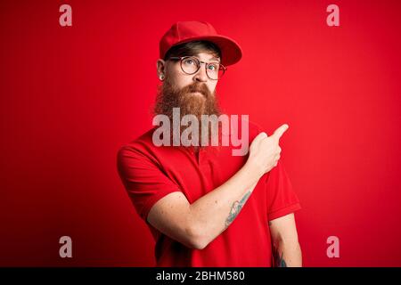 Giovane bel uomo di consegna che indossa occhiali e cappuccio rosso su sfondo isolato che punta con mano dito al lato mostrando pubblicità, grave Foto Stock