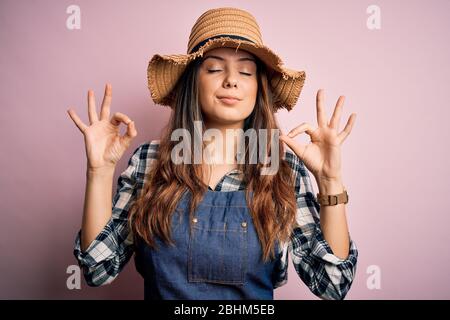 Giovane bella donna contadina bruna con grembiule e cappello su sfondo rosa rilassarsi e sorridere con gli occhi chiusi facendo un gesto di meditazione con il surf Foto Stock