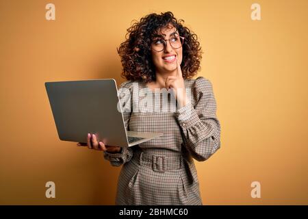 Bella donna d'affari araba che indossa occhiali che lavorano con il laptop su sfondo giallo faccia seria pensando alla domanda, idea molto confusa Foto Stock
