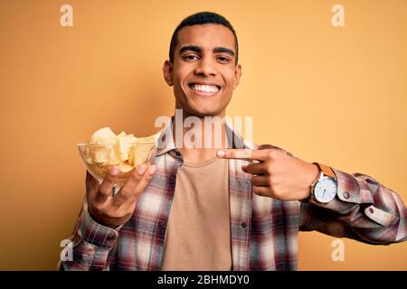 Giovane uomo africano amatone bello tenendo ciotola di patatine patatine su sfondo giallo molto felice puntando con mano e dito Foto Stock