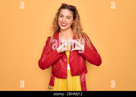 Giovane bella donna bionda pin-up con gli occhi blu indossando occhiali da sole rossi e giacca sorridente in amore mostrando il simbolo del cuore e la forma con le mani. Romant Foto Stock