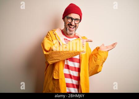 Uomo di età media che indossa occhiali e impermeabile in piedi su sfondo bianco isolato stupito e sorridente alla fotocamera mentre si presenta con la mano Foto Stock