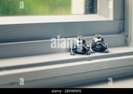 Due statuine del principe della rana coronate in argento sterling che siedono nel davanzale, concetto di colore. Foto Stock