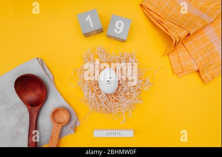 Uovo di Pasqua carino con un pennarello nero disegnato faccia in un nido di carta in carta pressata, cubi di legno grigio con la data di Pasqua aprile 19, napki cotone Foto Stock