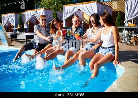 Gruppo di amici che si divertono a bordo piscina party clacking bicchieri con cocktail freschi spruzzi d'acqua presso la piscina nella soleggiata giornata estiva. Persone Foto Stock