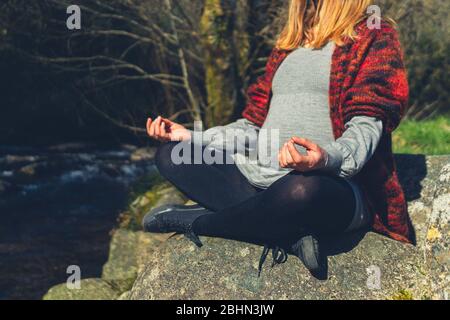 Una giovane donna incinta è seduta in una posa meditativa su una roccia vicino a un ruscello Foto Stock