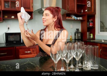 Sorridente bella giovane donna pulizia e bicchieri di vino al banco della cucina Foto Stock