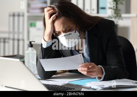 Triste donna esecutiva con maschera protettiva che lamenta guardando la ricevuta della banca in ufficio Foto Stock
