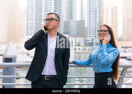 Il capo dell'uomo allegro sta avendo conversazione piacevole del telefono mobile con il cliente, mentre la sua segretaria femminile inoltre sta comunicando il telefono all'aperto sui grattacieli indietro Foto Stock