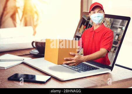 uomo in camicia rossa con maschera igienica, che tiene fuori l'ordine delle merci nel pacco dal computer portatile con sfondo del magazzino. ordine online, Foto Stock