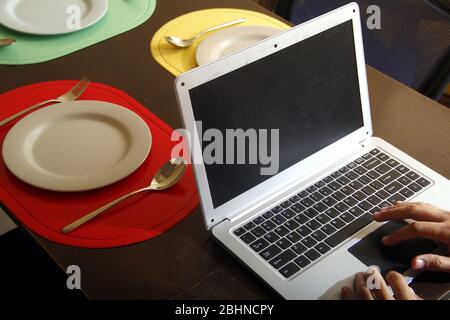Foto del computer portatile con le mani che scrivano su di esso, piatti e utensili su un tavolo da pranzo. Foto Stock
