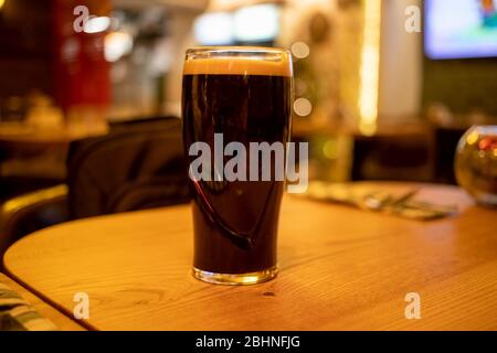 un bicchiere di birra scura stout con schiuma si erge su un tavolo di legno in un bar. Foto Stock