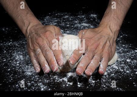 Le mani di un uomo impastano una palla di farina per fare il pane su una superficie nera piena di farina Foto Stock