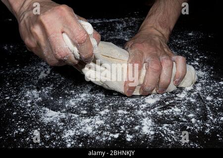 Le mani di un uomo separano un pezzo di pasta da una palla impastata con farina per fare il pane su una superficie nera piena di farina Foto Stock