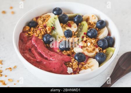 Ciotola per frullato di mirtillo rosa con frutta, bacche e granola in ciotola bianca su sfondo bianco. Concetto sano di cibo vegano. Foto Stock