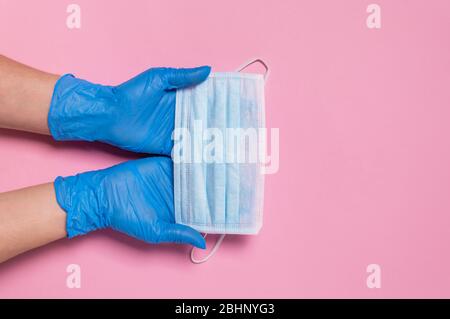 Protezione da coronavirus. Due mani in guanti monouso blu in lattice che tengono la maschera chirurgica medica su sfondo rosa. Misure igieniche per prevenire la diffusione Foto Stock