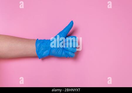 Mano del medico in guanto medico monouso blu che mostra i pollici su sfondo rosa. Gesto di approvazione o accettazione. Misure igieniche per prevenire le fuoriazioni Foto Stock