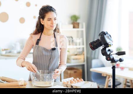 Buona-guardando giovane donna che indossa grembiule in piedi al tavolo in cucina moderna battendo le uova sulla macchina fotografica per il suo blog di cibo Foto Stock