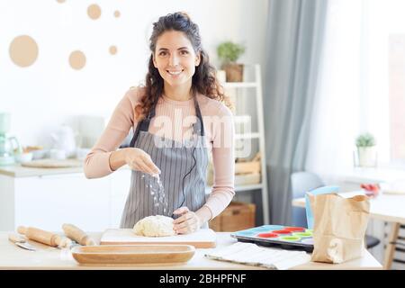 Buona-guardando donna caucasica che indossa grembiule in piedi al tavolo da cucina aggiungendo farina alla pasta guardando la macchina fotografica Foto Stock