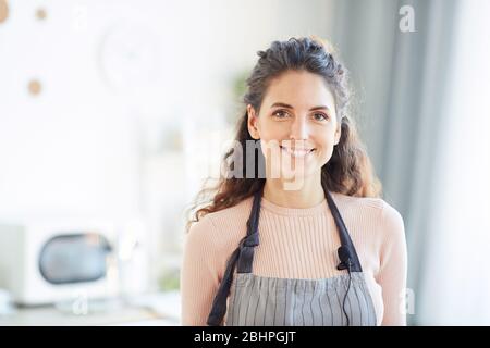 Donna caucasica elegante che indossa grembiule in piedi in cucina guardando la macchina fotografica sorridente, orizzontale petto su colpo Foto Stock