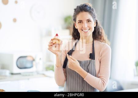 Orizzontale ritratto medio di bella giovane donna che indossa grembiule tenendo in casa dolce cupcake, guardando macchina fotografica sorridente Foto Stock