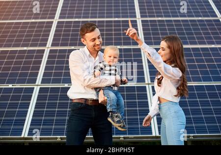 Il giovane padre sta tenendo il suo figlio piccolo, la madre sta indicando il cielo, il modulo PV grande è sullo sfondo, concetto di infanzia felice Foto Stock