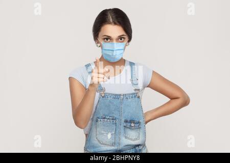 Ritratto di giovane donna bruna seria con maschera medica chirurgica in tuta denim in piedi guardando la macchina fotografica e avvertimento o scolante. Interni studi Foto Stock