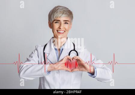 Concetto cardiologico. Medico femminile che mostra il cuore con le mani e cardiogramma su sfondo grigio, collage Foto Stock
