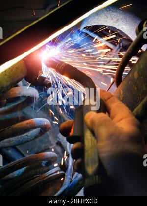 Lavori di saldatura presso una stazione di servizio auto: Primo piano di un meccanico che ripara le parti metalliche con un saldatore, colpo colorato Foto Stock