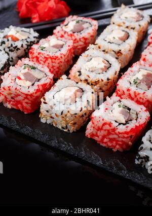 Panini di sushi uramaki con salmone, anguille affumicate, cetriolo, formaggio cremoso, sesamo, avocado e caviale su una tavola di pietra nera. Foto Stock