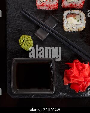 Cucina tradizionale Giapponese. Un set di sushi è servito con chopsticks e salsa di soia su ardesia nera, decorata con wasabi e zenzero. Foto Stock